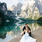 Bride and groom sitting by turquoise alpine lake with dramatic snow-capped mountain peaks reflected in water