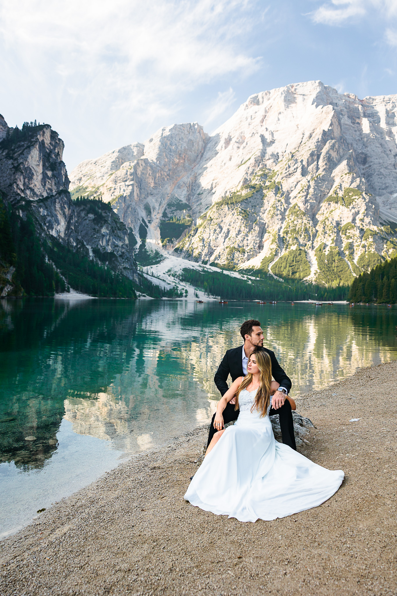 Bride and groom sitting by turquoise alpine lake with dramatic snow-capped mountain peaks reflected in water