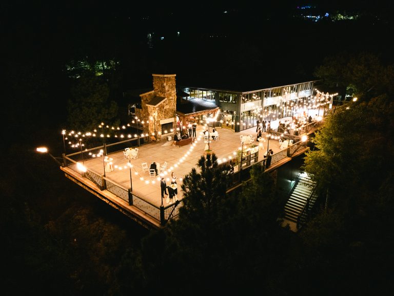 Nighttime aerial view of outdoor wedding venue with string lights, stone fireplace, and guests on illuminated terrace