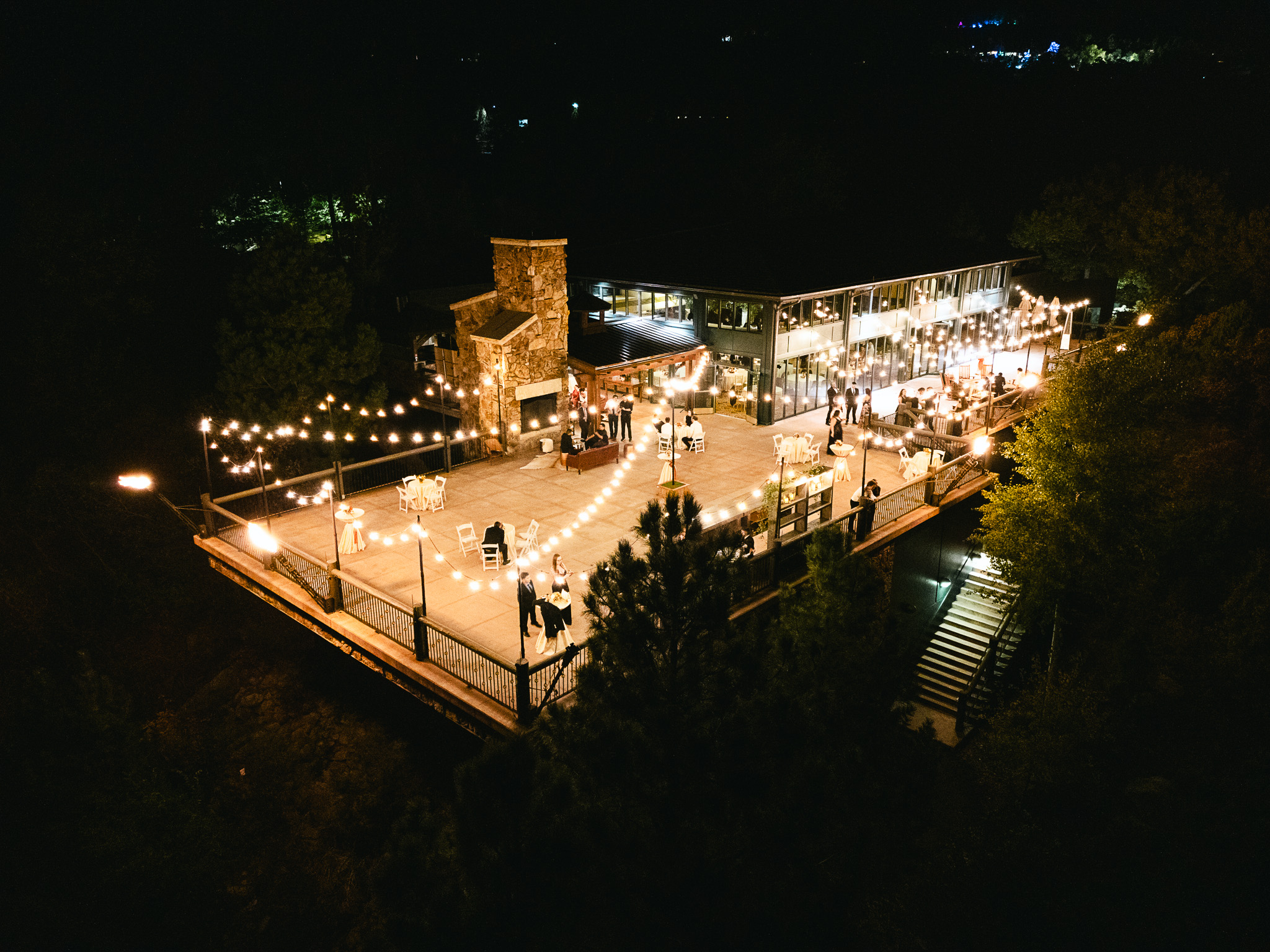 Nighttime aerial view of outdoor wedding venue with string lights, stone fireplace, and guests on illuminated terrace