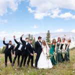 Bride and groom kissing in mountain meadow surrounded by groomsmen in black tuxedos and bridesmaids in emerald green gowns with Colorado peaks in background