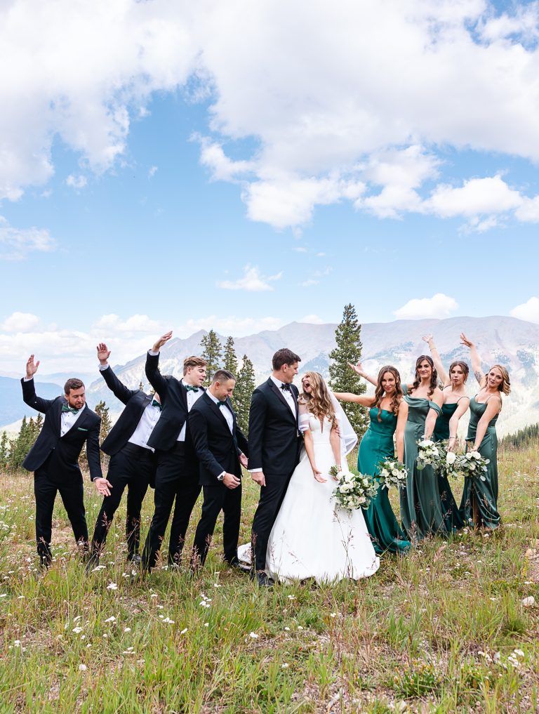 Bride and groom kissing in mountain meadow surrounded by groomsmen in black tuxedos and bridesmaids in emerald green gowns with Colorado peaks in background