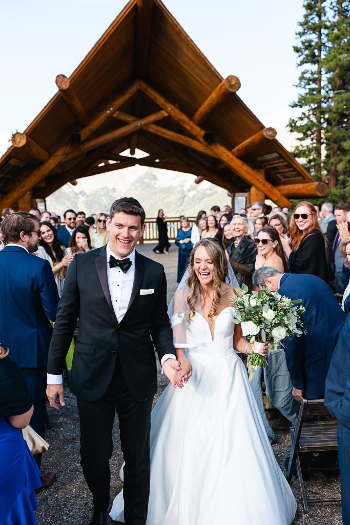 Newlyweds walking down the aisle hand-in-hand under a rustic log pavilion, surrounded by applauding guests at their Denver mountain wedding ceremony