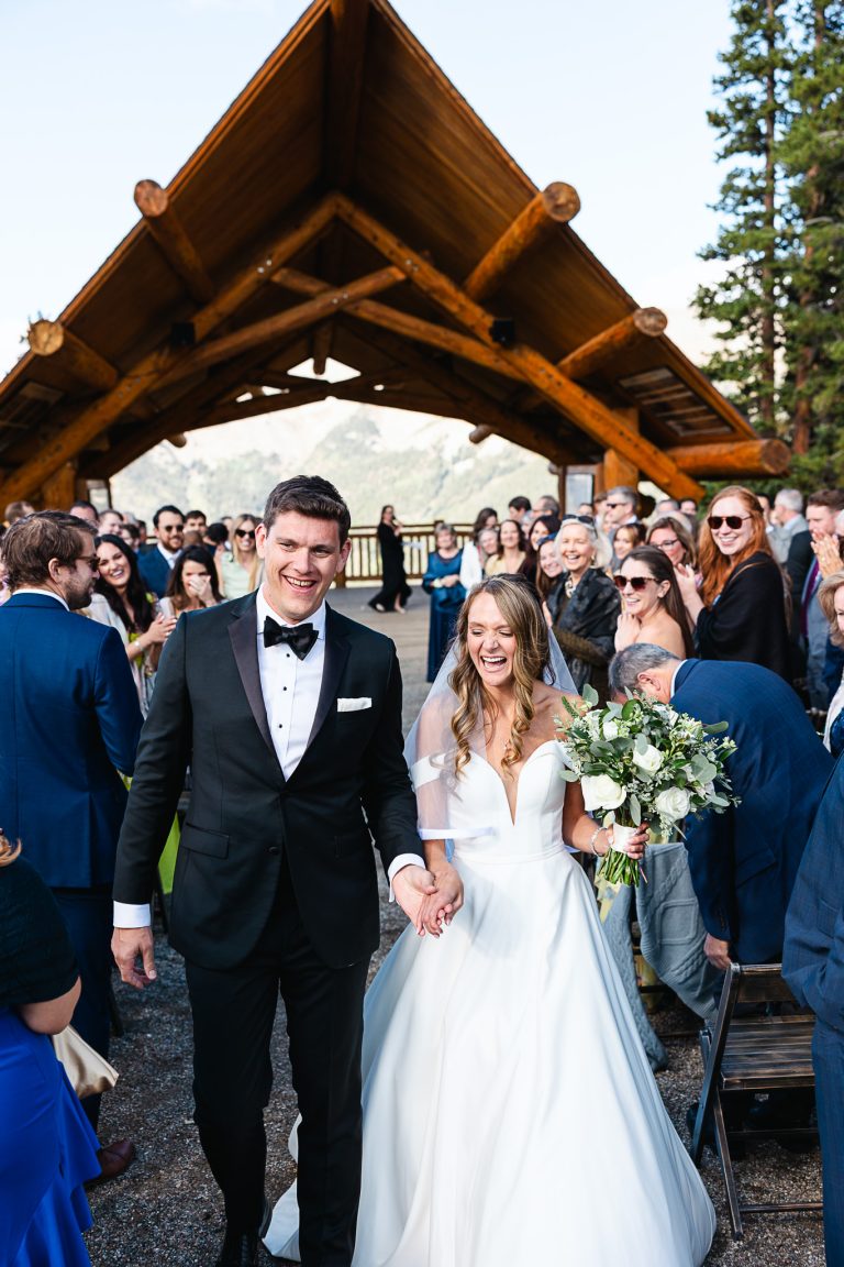 Newlyweds walking down the aisle hand-in-hand under a rustic log pavilion, surrounded by applauding guests at their Denver mountain wedding ceremony