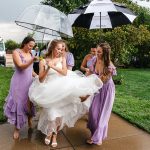 Bridesmaids in lavender dresses helping bride with flowing tulle gown under umbrellas on rainy wedding day