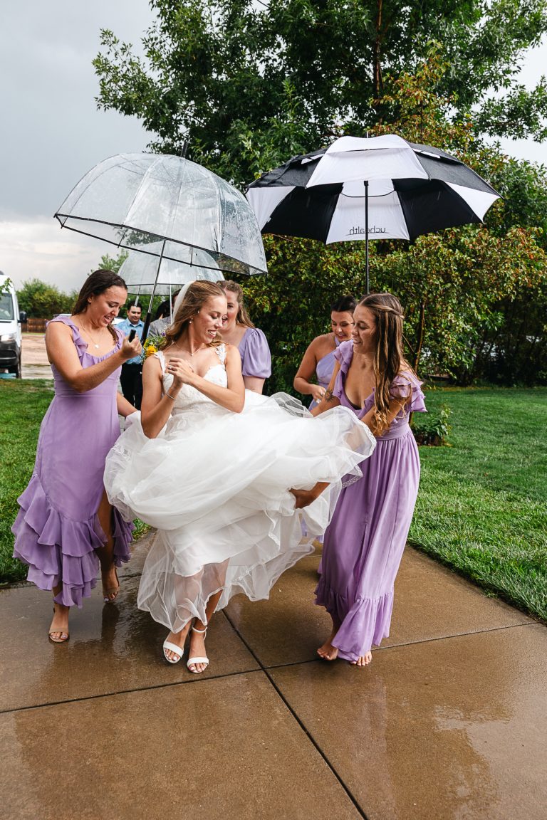 Bridesmaids in lavender dresses helping bride with flowing tulle gown under umbrellas on rainy wedding day