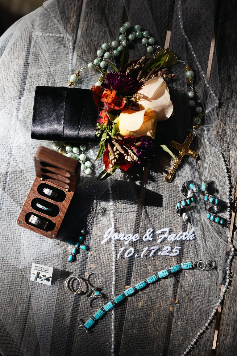 Overhead view of wedding preparation items including bridal bouquet with roses, jewelry, perfume, and accessories arranged on dark surface