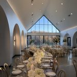 Elegant wedding reception venue with vaulted ceiling, arched windows, long tables adorned with white hydrangeas and candles beneath a dramatic triangular skylight