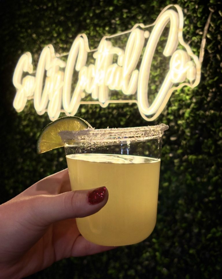 Margarita with lime wedge and salt rim held against greenery backdrop with "Cheers" neon sign
