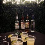 Margaritas with salted rims and lime garnishes displayed with premium liquor bottles under illuminated Sun City Cocktail Co. sign