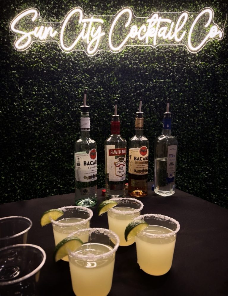 Margaritas with salted rims and lime garnishes displayed with premium liquor bottles under illuminated Sun City Cocktail Co. sign