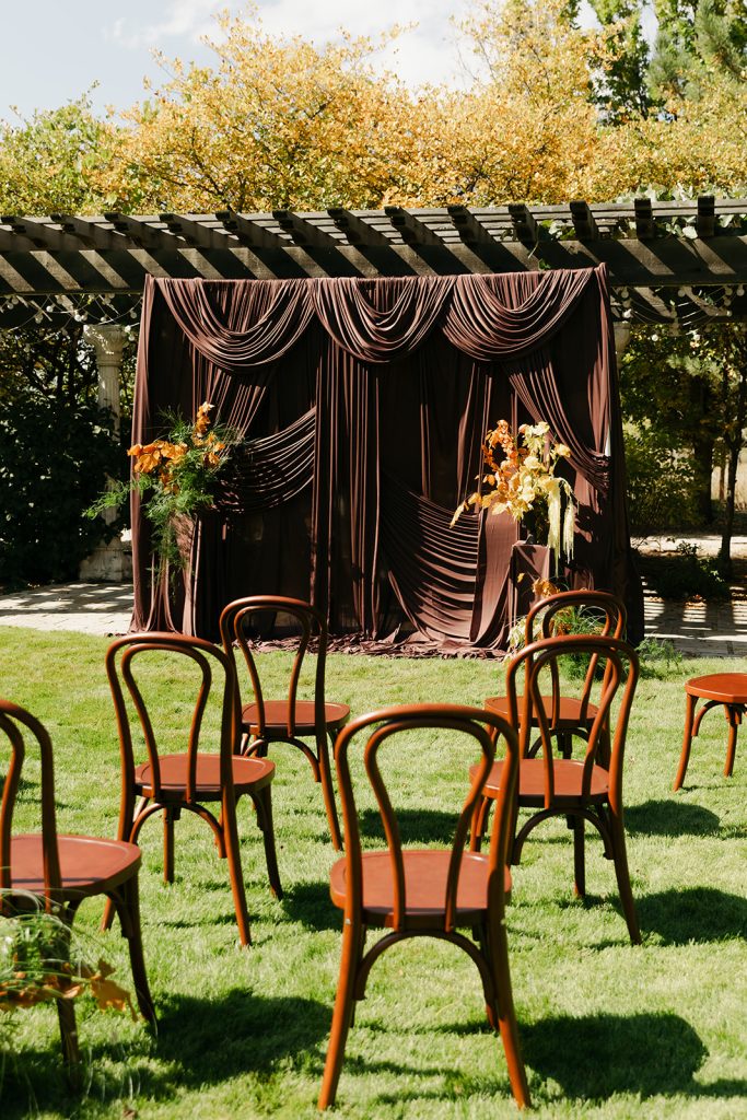 Outdoor wedding ceremony setup with wooden chairs, burgundy draped backdrop, and fall floral arrangements under pergola