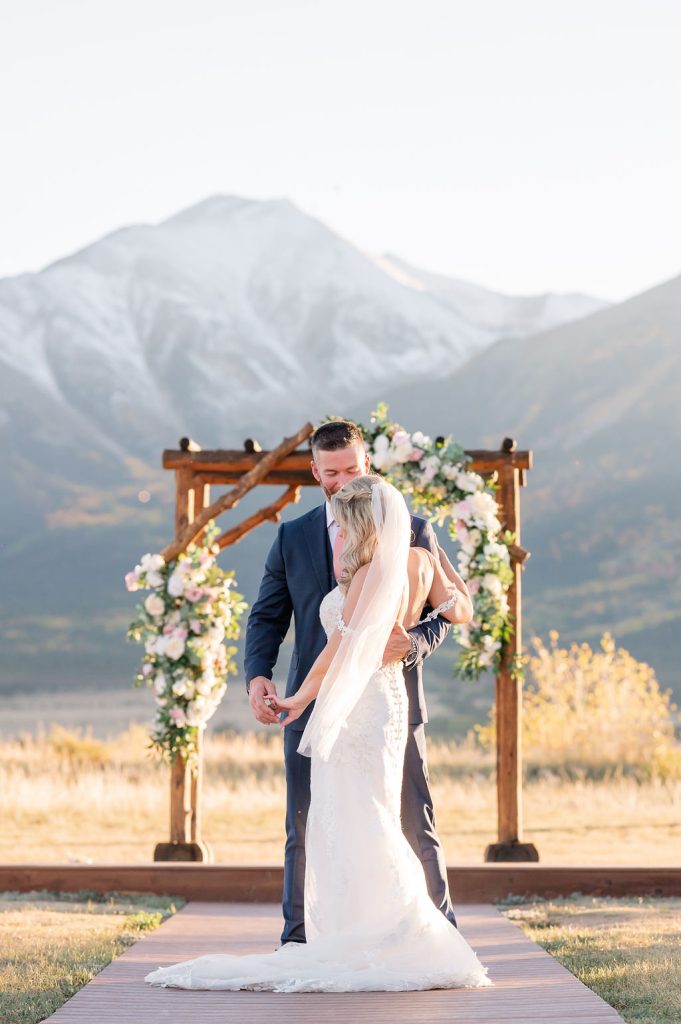 Newlyweds embrace under wooden ceremony arch with mountain backdrop in Colorado