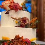 Three-tier white wedding cake decorated with fall flowers in burgundy and orange tones