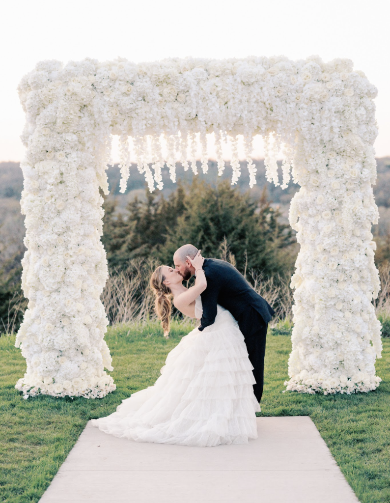 Bride and groom kiss beneath elaborate white floral arch with cascading blooms during outdoor ceremony