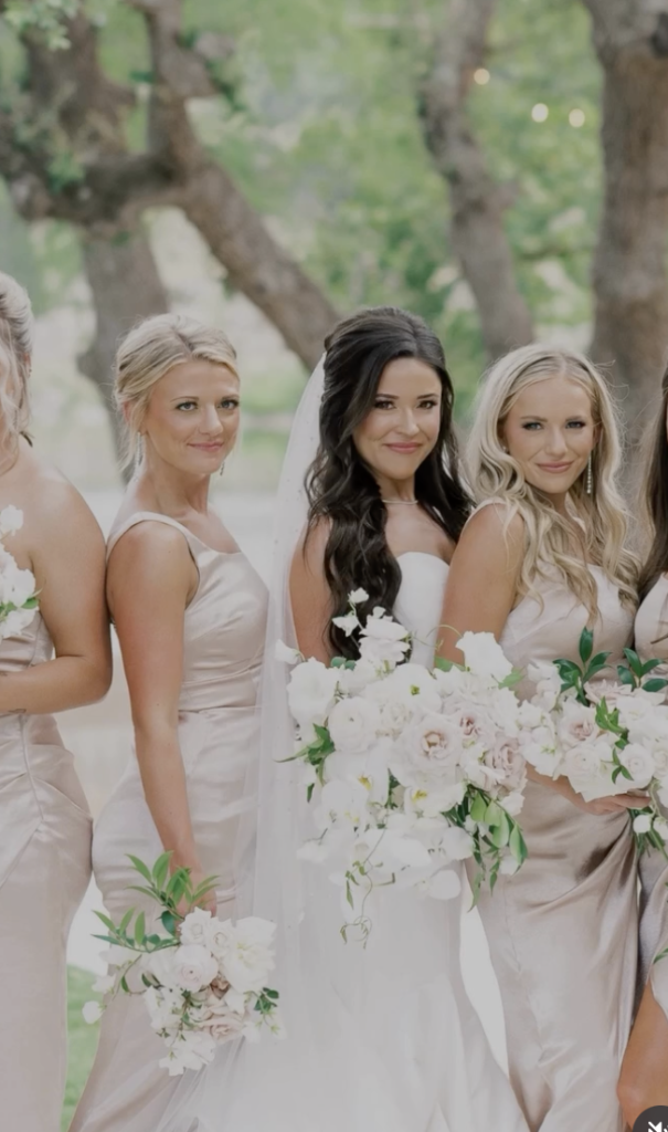 Bride and bridesmaids in neutral gowns hold white bouquets under oak trees