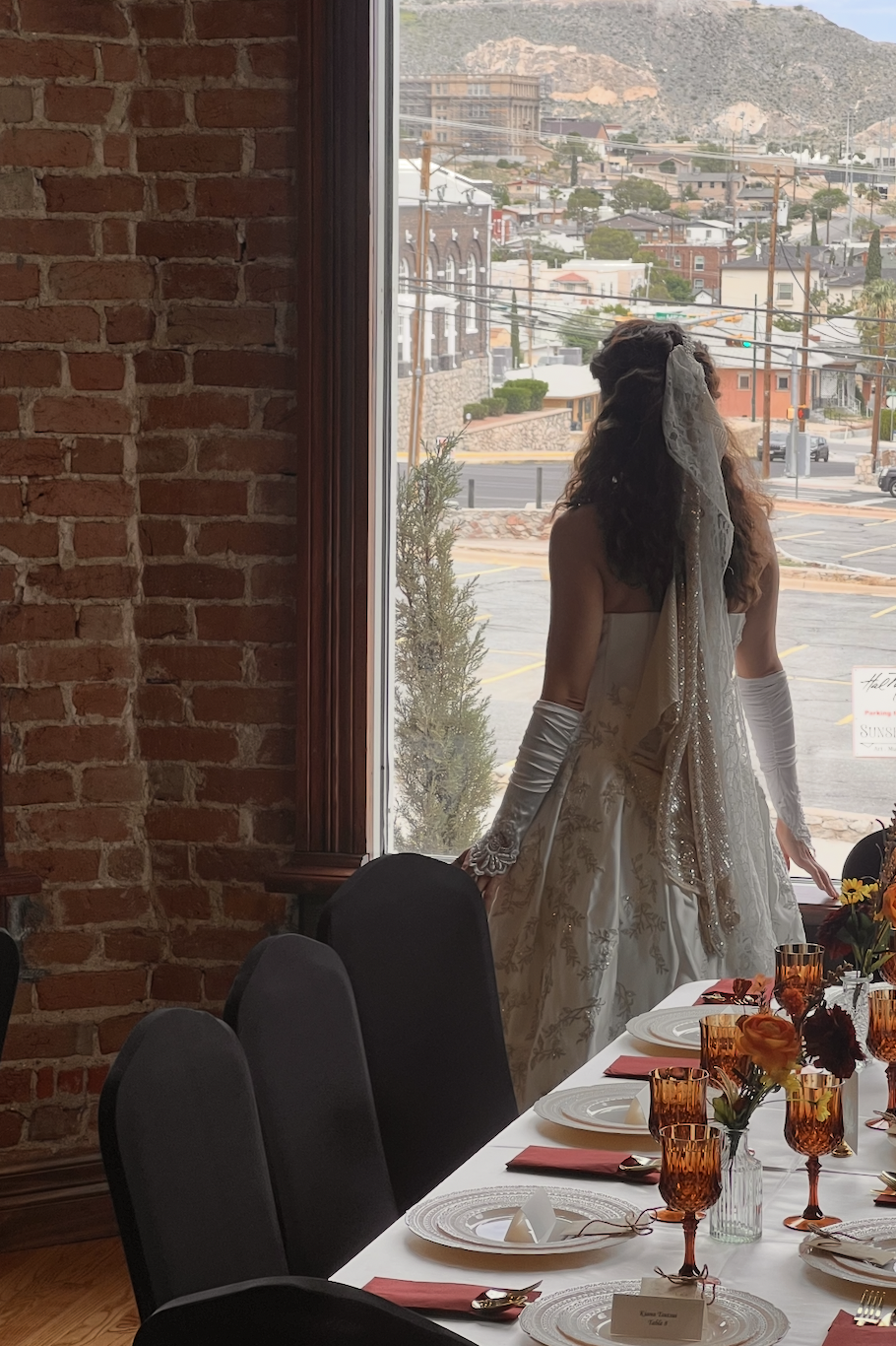 Bride with veil and gloves admiring mountain views beside formal dining table with amber glassware and black velvet chairs
