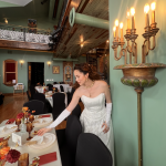 Bride in white gown arranging table settings in elegant reception hall with vaulted wood ceiling and ornate gold candelabra
