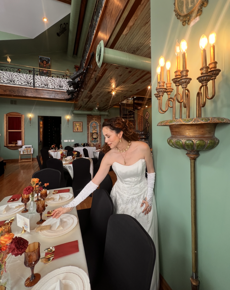 Bride in white gown arranging table settings in elegant reception hall with vaulted wood ceiling and ornate gold candelabra