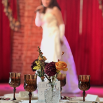 Wedding reception table setting with amber glassware, burgundy and orange floral centerpiece, and red velvet draping