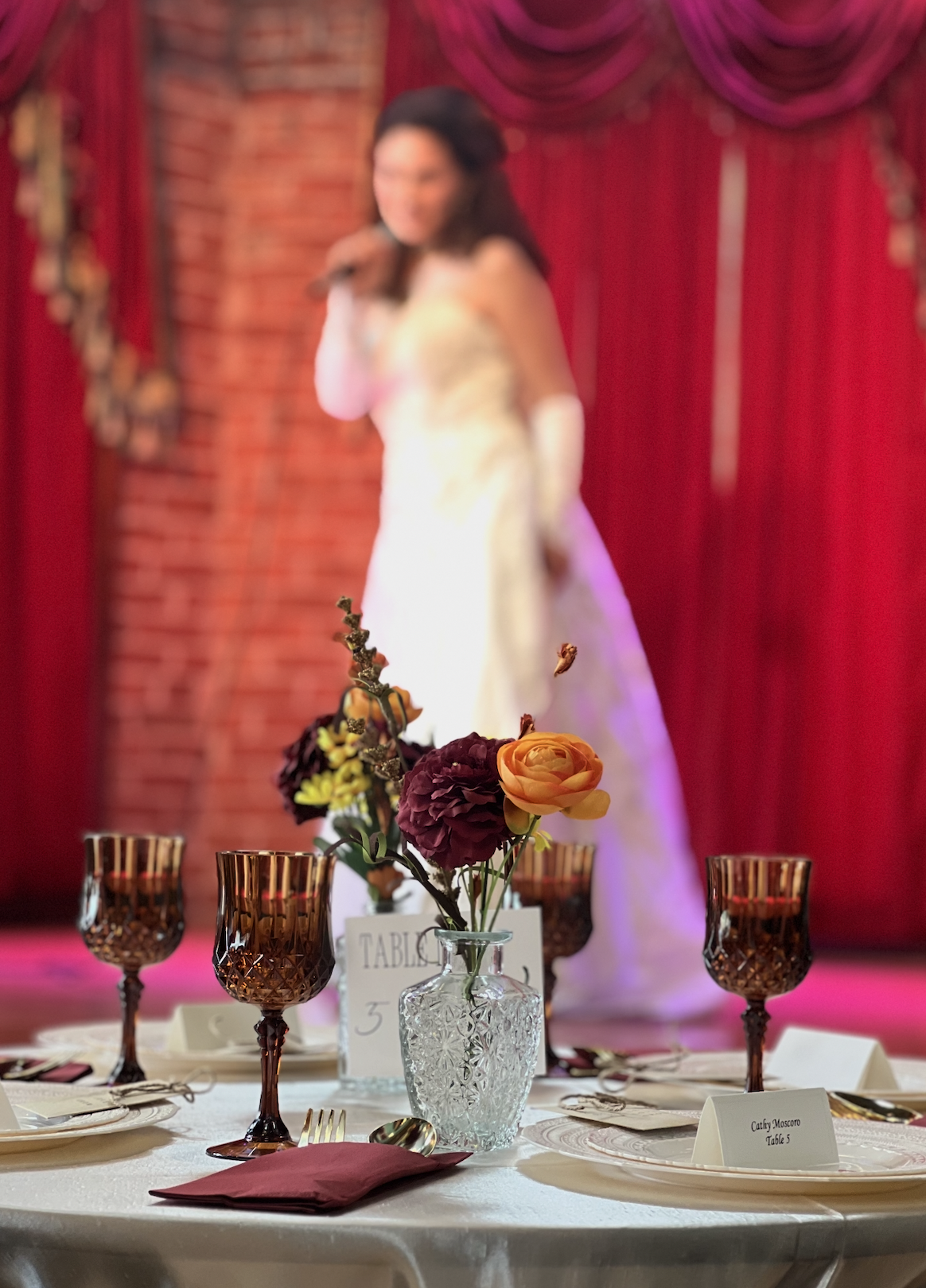 Wedding reception table setting with amber glassware, burgundy and orange floral centerpiece, and red velvet draping