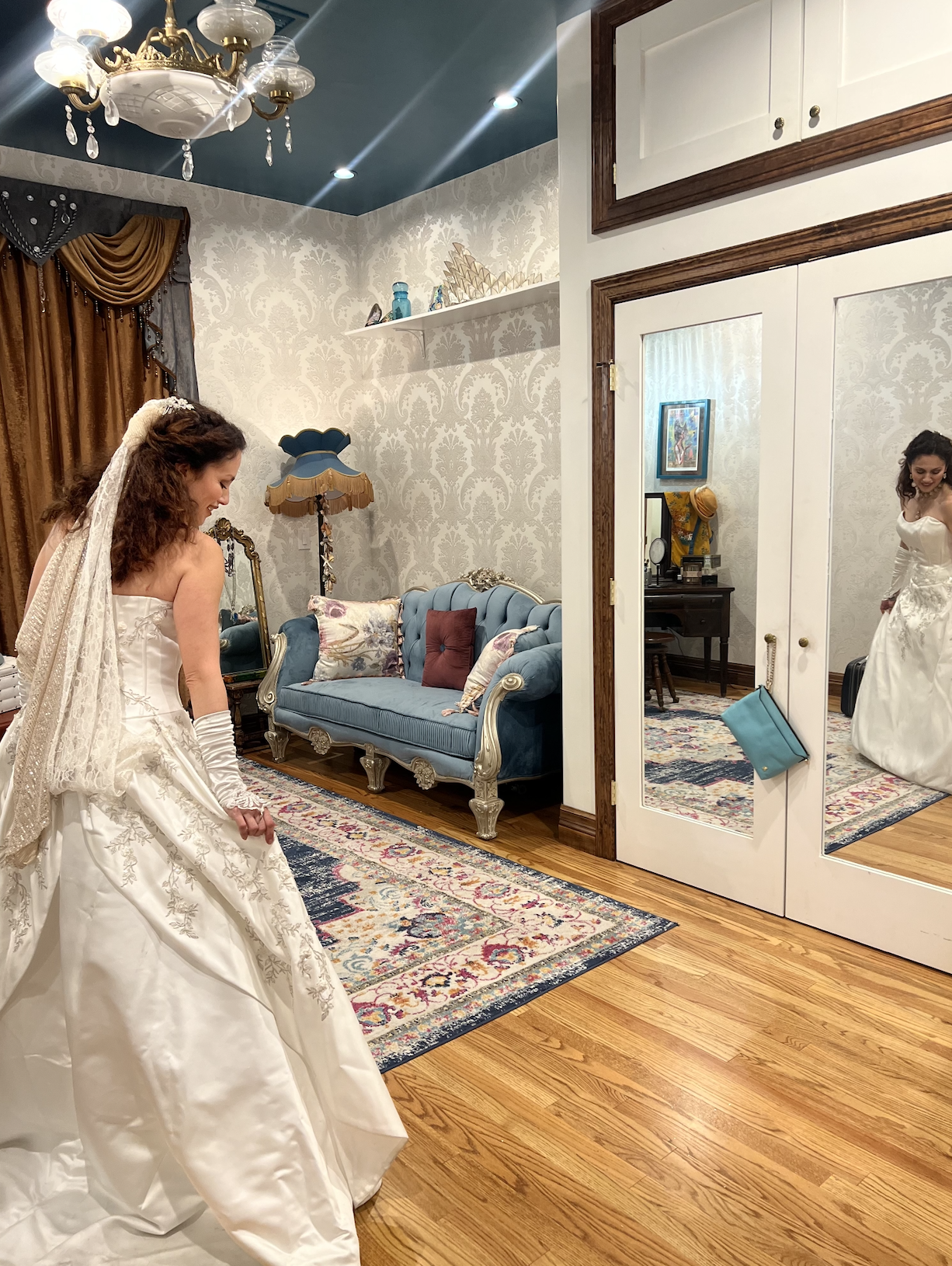 Bride in lace wedding gown admiring her reflection in elegant bridal suite with vintage blue settee and damask wallpaper