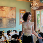 Guest in beaded formal attire standing beside reception table with fall-colored florals, exposed brick walls, and crystal chandelier