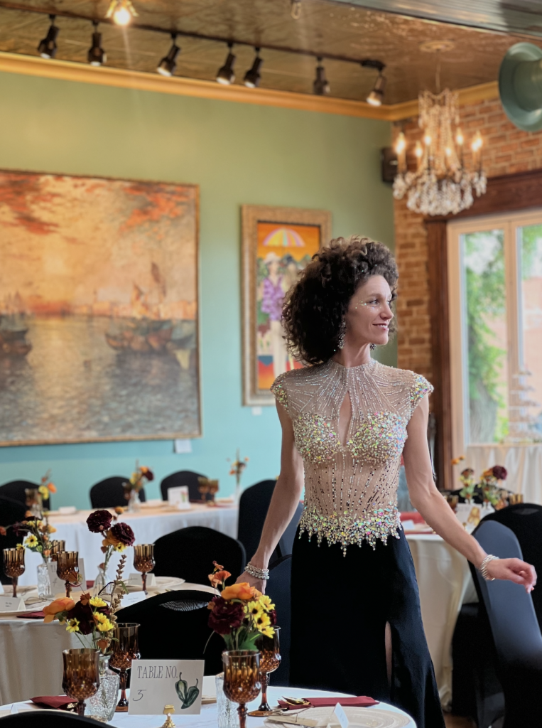 Guest in beaded formal attire standing beside reception table with fall-colored florals, exposed brick walls, and crystal chandelier