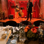 Jazz duo performing on stage with red velvet backdrop, view from formal dining table with floral centerpiece