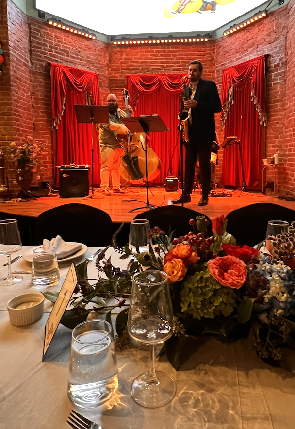 Jazz duo performing on stage with red velvet backdrop, view from formal dining table with floral centerpiece