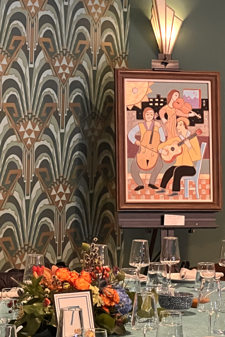 Art Deco-styled reception table with colorful floral centerpiece, elegant glassware, and vintage jazz musicians artwork on green wall