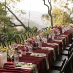 Long outdoor reception table with burgundy runners, pink blooms, and black chairs overlooking mountain vista