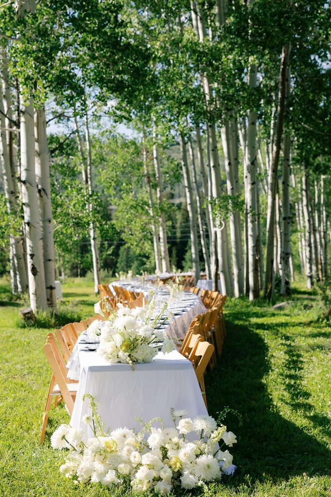 Outdoor wedding reception table with white linens and lush white floral arrangements among aspen trees