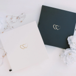 Marriage license folders in cream and forest green with interlocking heart logos, displayed with pearl jewelry and white flowers