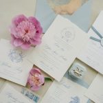 Coastal-themed wedding invitation suite with blue script typography, pink peonies, seahorse motifs, and vintage postage stamps