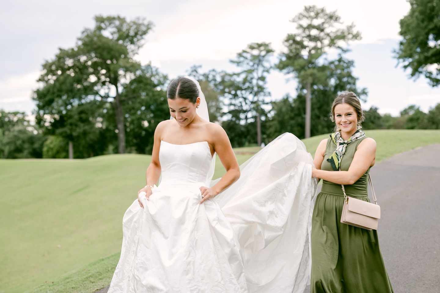 Bride in strapless wedding gown with bridesmaid in green dress helping arrange the dress train on golf course