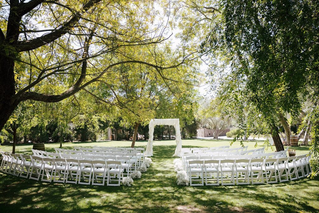 Outdoor wedding ceremony setup with white folding chairs arranged in curved rows under tall trees, featuring white ceremony arch on manicured lawn