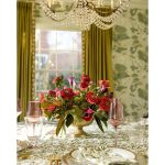Elegant red and pink floral centerpiece in gold compote vase beneath crystal chandelier on patterned tablecloth