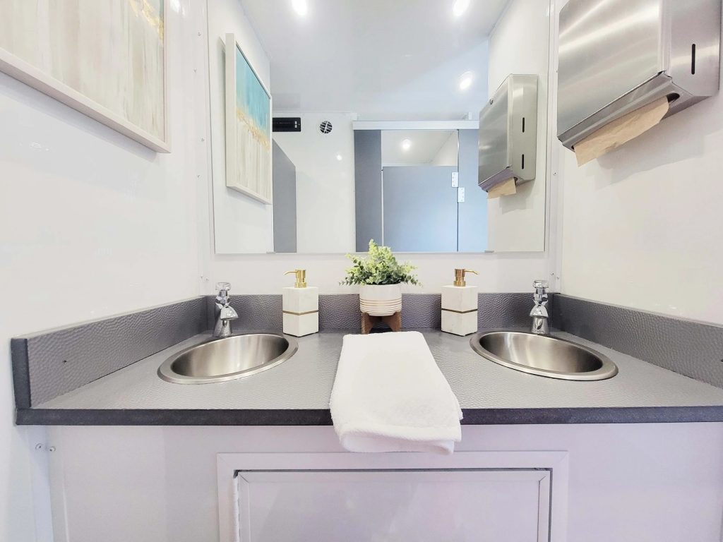Upscale portable restroom suite interior featuring dual stainless steel sinks, elegant white soap dispensers with gold pumps, fresh greenery centerpiece, and modern white cabinetry