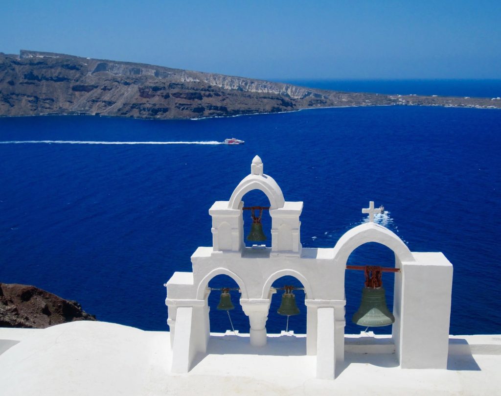 White church bell tower with brass bells overlooking deep blue Aegean Sea and volcanic cliffs of Santorini