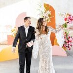 Bride and groom celebrating with champagne in modern studio with colorful geometric backdrop and floral arrangements