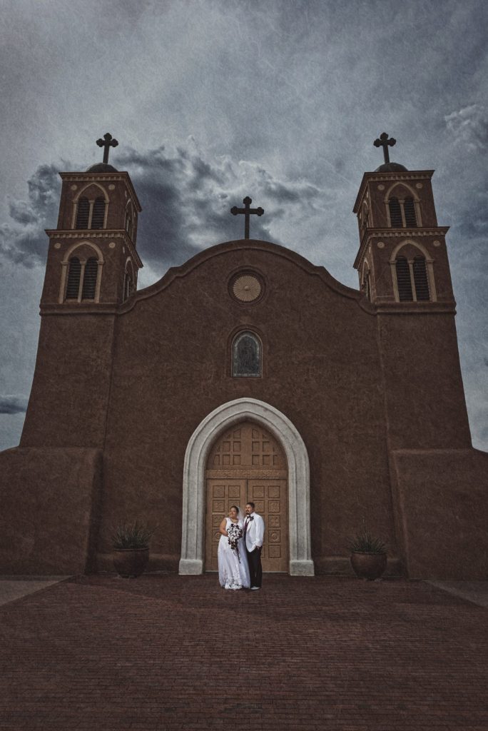 Bride and groom standing in doorway of historic adobe church with twin bell towers under dramatic cloudy sky