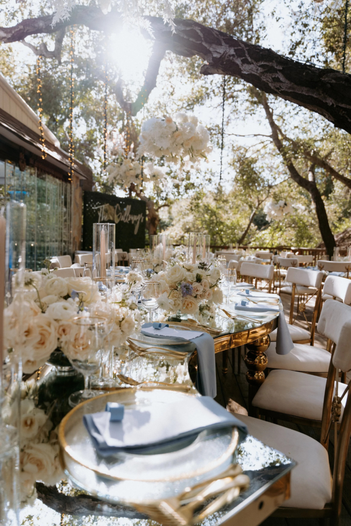 Elegant outdoor wedding reception with gold chiavari chairs, blue and white place settings, and tall white floral centerpieces under trees