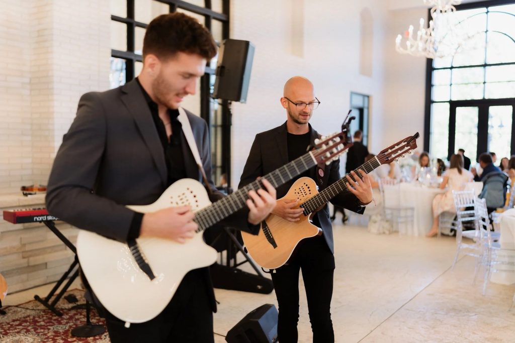 Two classical guitarists in black suits perform at an elegant wedding cocktail hour in a white industrial venue