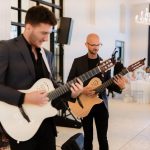 Two acoustic guitarists in black suits performing at elegant white wedding reception venue