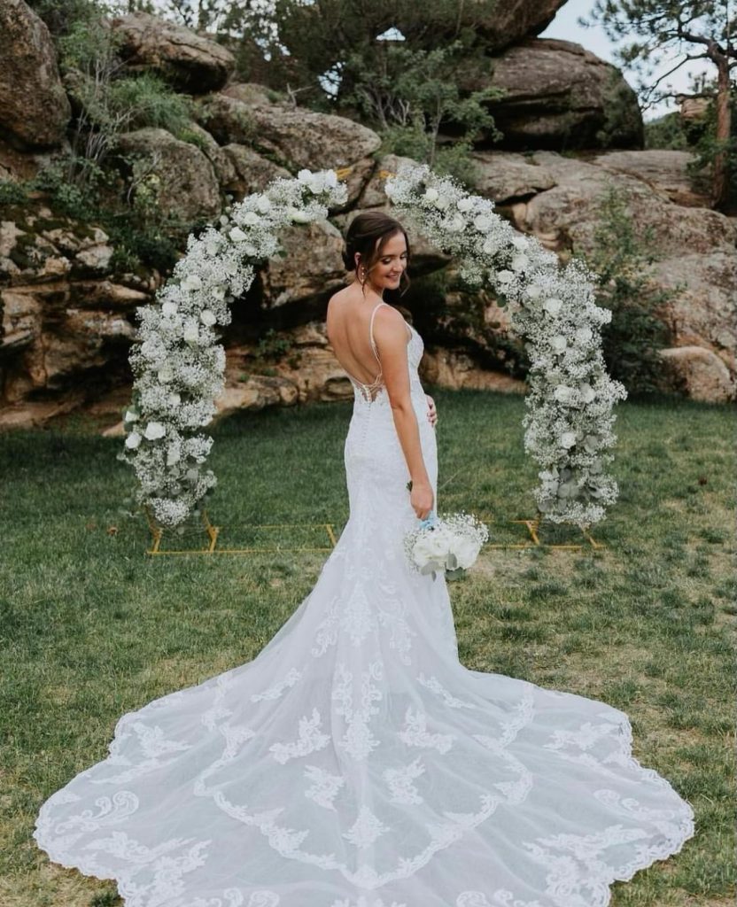 Bride in lace gown with flowing train standing before white floral arch at outdoor Colorado rock garden venue