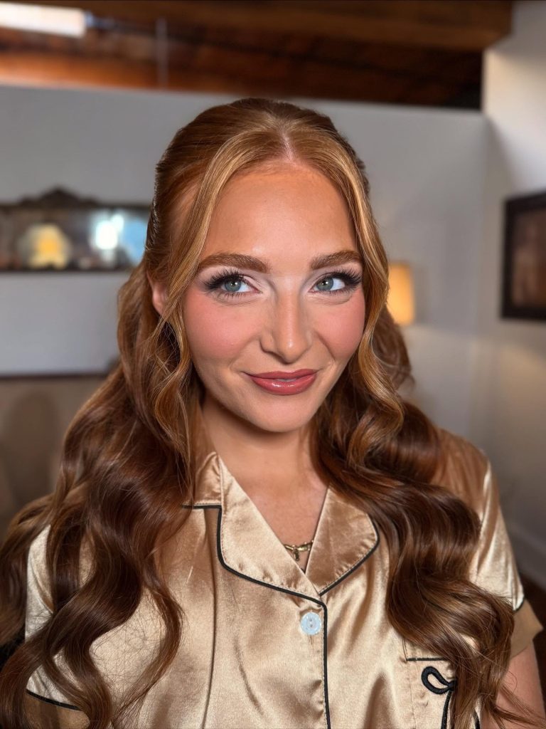 Bride with long wavy auburn hair styled half-up, wearing natural bridal makeup with defined lashes