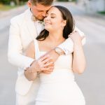 Bride and groom in elegant white attire embrace on tree-lined street in St. Louis