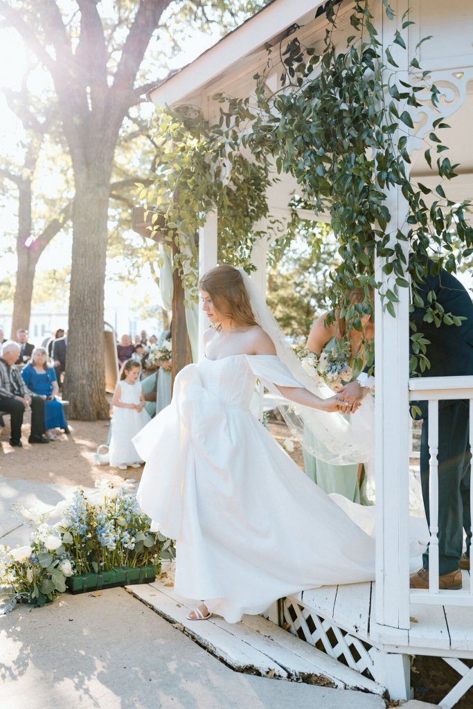 Bride's dress billowing as she enters outdoor ceremony through white gazebo adorned with eucalyptus
