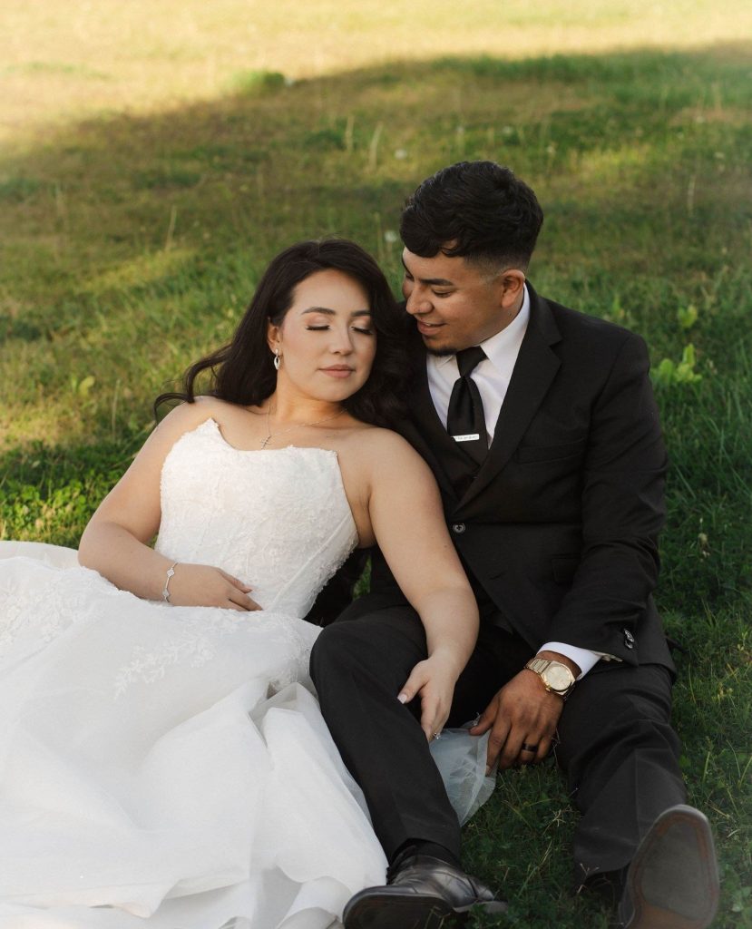 Newlywed couple sitting intimately on grass hillside during golden hour, bride in white gown and groom in black tuxedo